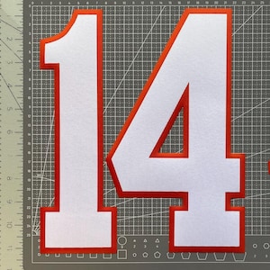May include: White felt number 14 with a red border, displayed on a gray cutting mat. The numbers are large and bold, suitable for crafts or decorations. The image is a close-up, highlighting the clean lines and color contrast.