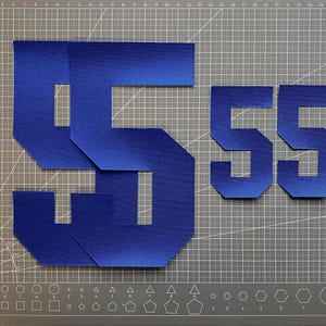 May include: Blue felt number 55 cutouts. The numbers are in a bold, angular font. The image shows a large number 5, a smaller number 5, and two additional number 5 cutouts. The cutouts are on a grey cutting mat.