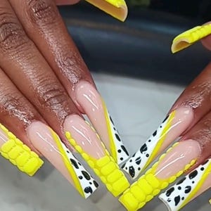 May include: Long, square-shaped artificial nails featuring a combination of bright yellow 3D textured raised patterns, a nude base, and white tips adorned with black cow-print spots. The design includes diagonal yellow lines separating the patterns.