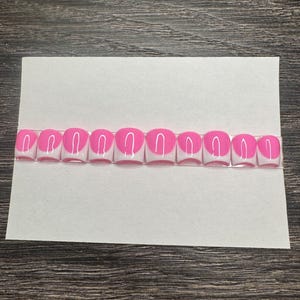 May include: A set of ten artificial nails with a bright pink half-moon design on a white base. The nails are arranged in a row on a white card, ready for application. The background is a dark wood grain.