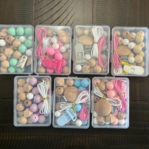 May include: Six clear plastic containers filled with wooden beads and clips. The beads are in various colors, including pink, blue, green, and brown. The containers are arranged in two rows of three.