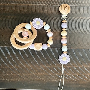 May include: A wooden baby teether with a flower design and a matching wooden and beaded pacifier clip with a flower charm. The clip has a wooden tag with the letter "R" engraved on it.