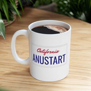Peut inclure: Une tasse à café en céramique blanche avec une poignée noire. La tasse est décorée d'un motif rouge et bleu qui dit "California ANUSTART".