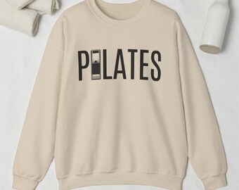 May include: A cream-coloured Pilates sweatshirt featuring the word "PILATES" in black, with the letter "I" replaced by a black and white Pilates reformer illustration. The sweatshirt is made of a soft material.