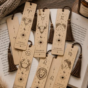 Puede incluir: Colección de marcapáginas rectangulares con ilustraciones de signos del zodiaco y borlas a juego. Cada marcapáginas presenta un signo diferente, como Aries, Tauro y Géminis, con símbolos y texto asociados. Los marcapáginas están hechos de un material marrón claro.