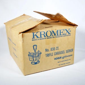 Könnte beinhalten: Eine Vintage-Kartonbox mit dem blauen Text "KROMEX" und "TRIPLE CAROUSEL SERVER". Die Box ist leicht beschädigt, mit einer offenen Klappe oben. Die Box hat auch eine Grafik des Produkts.