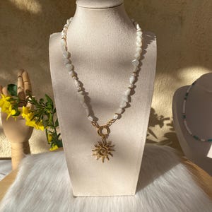 Peut inclure: Un collier de perles blanches avec un pendentif soleil doré, présenté sur un mannequin. Le collier est composé de perles blanches irrégulières et de petits accents dorés. Le pendentif soleil est orné de rayons tourbillonnants.