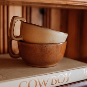 May include: Two stacked ceramic mugs with a smooth, rounded shape and square-angled handles. The top mug is a light cream color, while the bottom mug is a darker, earthy brown. Both mugs feature a subtle, speckled texture across their surfaces.