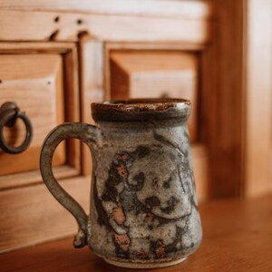 May include: A handcrafted ceramic mug with a textured, mottled surface in shades of gray, brown, and muted blue. The mug features a thick, rounded rim, a sturdy curved handle, and a tapered cylindrical body with visible earthy, rustic glaze variations.