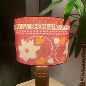 May include: A table lamp with a wooden base and a red lampshade. The lampshade has a floral pattern with white and yellow accents and text. The lamp is on a small wooden table. The lamp is a decorative item.