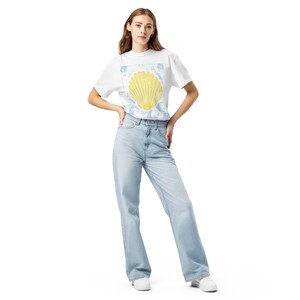 Peut inclure: Une femme porte un t-shirt blanc avec un motif de coquillage jaune et un jean bleu clair. Le t-shirt a un col rond et des manches courtes. Le jean est taille haute et à jambes larges. Elle porte des baskets blanches.