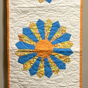 May include: A quilted table runner featuring three large, stylized flower designs. Each flower has a bright orange center and blue petals with a yellow and green patterned fabric. The runner is bordered in orange and has a white background.