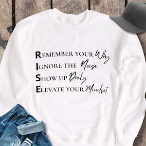 Peut inclure: Sweat-shirt blanc avec l'acronyme RISE et les mots "Remember Your Why", "Ignore The Noise", "Show Up Daily", et "Elevate Your Mindset" imprimés en noir.