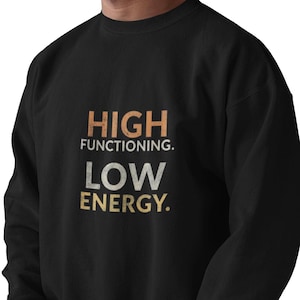 Peut inclure: Sweat-shirt noir avec l'inscription "HIGH FUNCTIONING. LOW ENERGY." en orange et jaune. Le sweat-shirt est de style col rond et est porté par une personne.