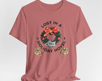 May include: Dusty rose t-shirt with a graphic featuring a Christmas wreath with a red bow and the text "Lost in a Holiday Novel." Two books are on either side of the wreath. The words "Merry Christmas" are in the center of the wreath.