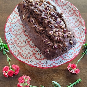 May include: A loaf of chocolate banana bread topped with pecan nuts, sitting on a red and white patterned plate.