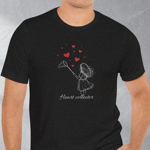 May include: Black t-shirt with a white graphic of a girl catching red hearts with a net. The text "Heart collector" is below the graphic. The shirt is worn by a person.