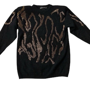 Puede incluir: Suéter negro con mangas largas y cuello redondo. El suéter presenta un patrón de lentejuelas de color bronce en un diseño abstracto y fluido en la parte delantera. La prenda está hecha de un material suave.