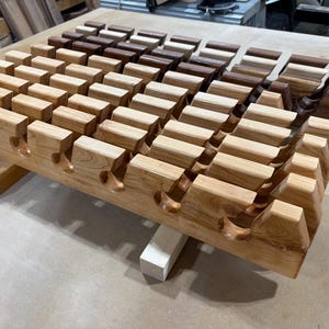 Puede incluir: Un soporte de madera con m&uacute;ltiples filas de ranuras talladas dise&ntilde;adas para sostener objetos. El soporte est&aacute; construido con madera de color claro en la parte delantera y cambia a tonos de madera marr&oacute;n oscuro y rico hacia la parte trasera.