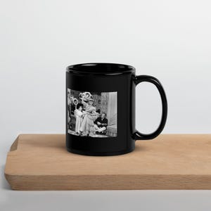 May include: Black ceramic mug with a black and white image of a vintage film scene. The image depicts people in period costume, possibly from a play or film. The mug is placed on a wooden surface.