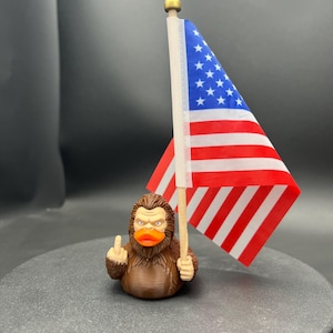 May include: A brown rubber duck figurine with a Bigfoot design, holding a small American flag and giving the middle finger. The flag has red and white stripes and a blue field with white stars. The duck has an orange beak.