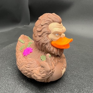 Puede incluir: Una figura de pato de goma marr&oacute;n y texturizada inspirada en un Sasquatch, con un pico naranja brillante y una cara de color canela claro. La figura muestra un dedo medio levantado en un ala, hojas verdes y una flor rosa brillante en la espalda.