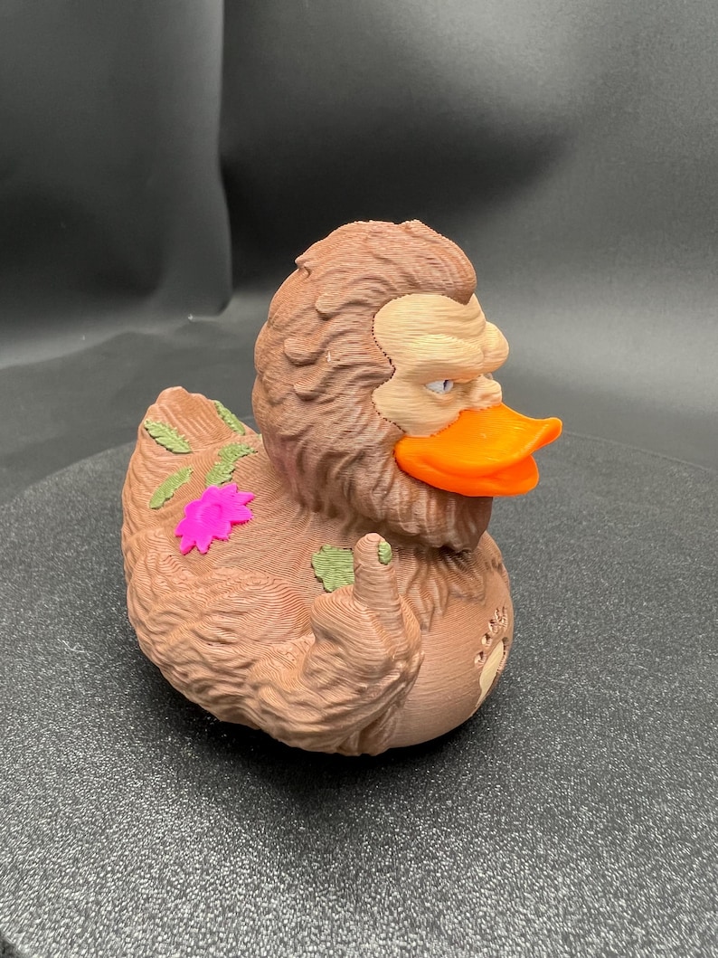 Puede incluir: Una figura de pato de goma marr&oacute;n y texturizada inspirada en un Sasquatch, con un pico naranja brillante y una cara de color canela claro. La figura muestra un dedo medio levantado en un ala, hojas verdes y una flor rosa brillante en la espalda.
