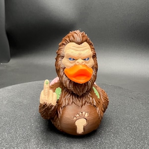 Puede incluir: Una figura de criatura marr&oacute;n y peluda con un pico de pato naranja brillante, ojos azules y un emblema de huella de pie de color claro en el pecho. La figura tiene un brazo levantado mostrando el dedo medio y detalles de hojas verdes.