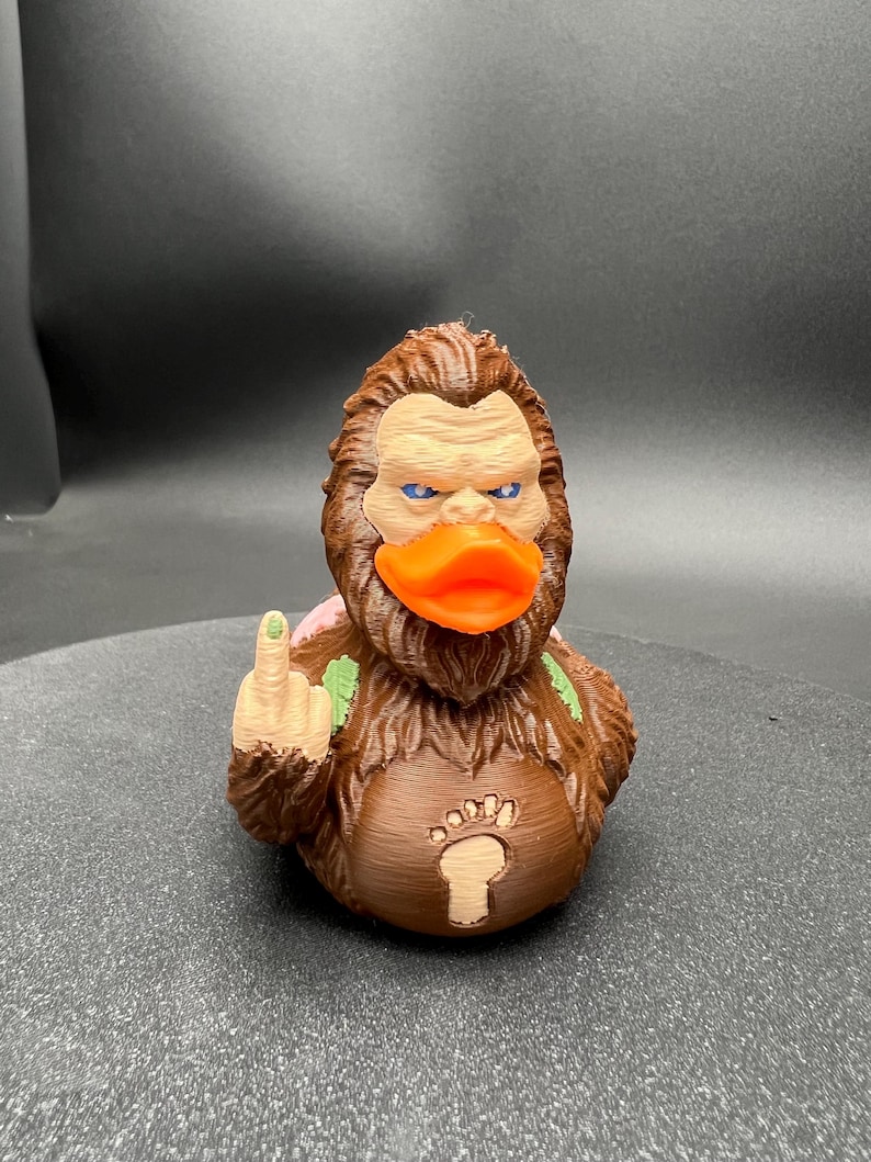 Puede incluir: Una figura de criatura marr&oacute;n y peluda con un pico de pato naranja brillante, ojos azules y un emblema de huella de pie de color claro en el pecho. La figura tiene un brazo levantado mostrando el dedo medio y detalles de hojas verdes.