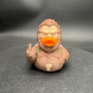 Puede incluir: Una figura de pato de goma texturizada de color marr&oacute;n inspirada en Bigfoot con un pico naranja brillante. La figura presenta una cara de color marr&oacute;n claro, un emblema de huella en el pecho y un dedo medio levantado en su ala derecha.