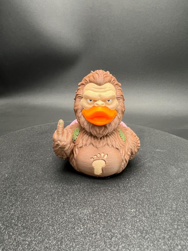 Puede incluir: Una figura de pato de goma texturizada de color marr&oacute;n inspirada en Bigfoot con un pico naranja brillante. La figura presenta una cara de color marr&oacute;n claro, un emblema de huella en el pecho y un dedo medio levantado en su ala derecha.