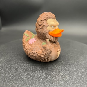 Puede incluir: Una figura de pato de goma texturizada de color marr&oacute;n con apariencia de Sasquatch, que presenta un pico naranja brillante, una cara de color tostado claro y un detalle de flor rosa en la espalda. La figura tiene un dedo medio levantado y un icono de huella.