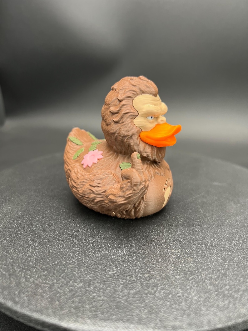 Puede incluir: Una figura de pato de goma texturizada de color marr&oacute;n con apariencia de Sasquatch, que presenta un pico naranja brillante, una cara de color tostado claro y un detalle de flor rosa en la espalda. La figura tiene un dedo medio levantado y un icono de huella.