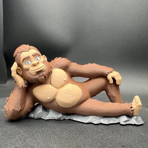 Op de afbeelding: Bruin en tan beeldje van een ontspannen Bigfoot, liggend op een grijze stenen basis. De figuur heeft blauwe ogen en poseert met een arm achter het hoofd. De vacht is getextureerd en het algehele ontwerp is cartoonachtig.