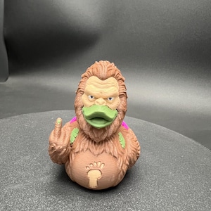 Puede incluir: Una figura de pato de goma texturizada de color marr&oacute;n, similar a un simio, con pico verde, peque&ntilde;as alas moradas en la espalda y un emblema de huella en el pecho. La figura muestra un dedo medio levantado en el lado derecho con un punto verde.
