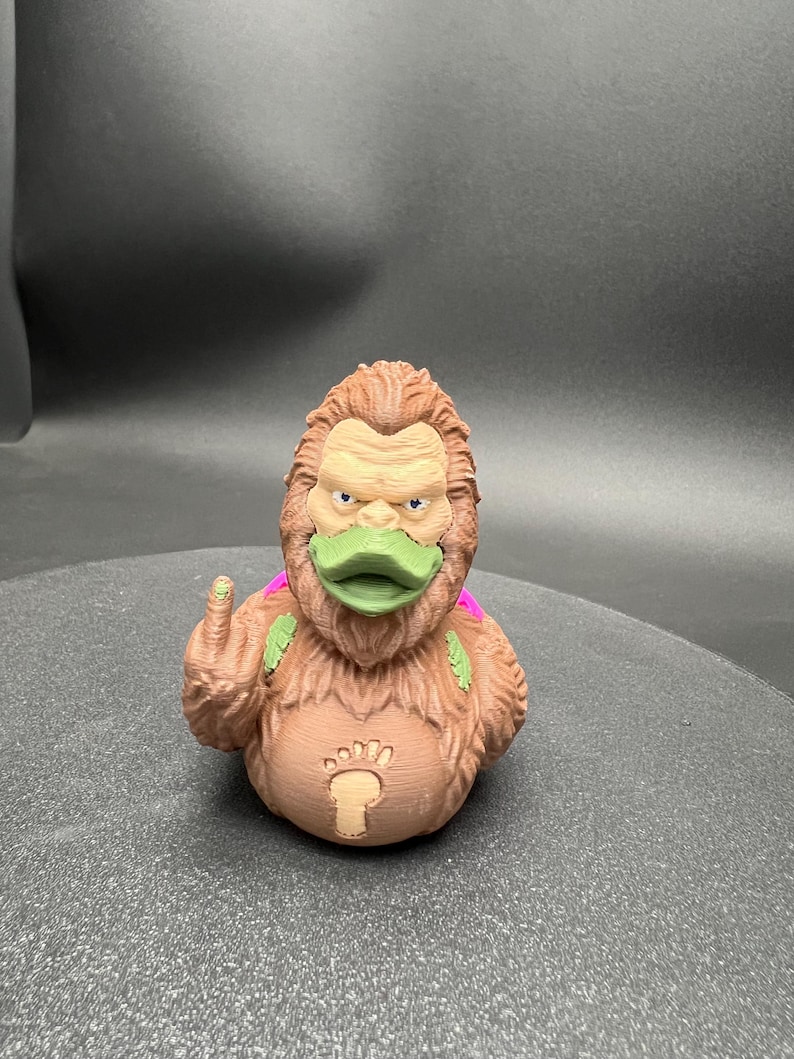 Puede incluir: Una figura de pato de goma texturizada de color marr&oacute;n, similar a un simio, con pico verde, peque&ntilde;as alas moradas en la espalda y un emblema de huella en el pecho. La figura muestra un dedo medio levantado en el lado derecho con un punto verde.