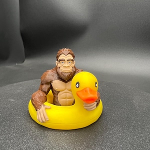 Könnte beinhalten: Eine braune, muskulöse Bigfoot-Figur mit fellartiger Textur, die in einem leuchtend gelben Gummienten-Schwimmring sitzt. Die Ente hat einen leuchtend orangefarbenen Schnabel und schwarze Augen, während die Bigfoot-Figur hellbraune Brustdetails aufweist.
