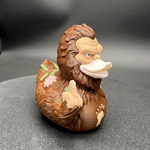 Puede incluir: Una figura con forma de pato, marr&oacute;n y peluda, con cara de color tostado claro y pico blanco. La figura tiene una mano levantada haciendo un gesto con el dedo coraz&oacute;n, un emblema de huella en el pecho y peque&ntilde;as flores rosas y hojas verdes.