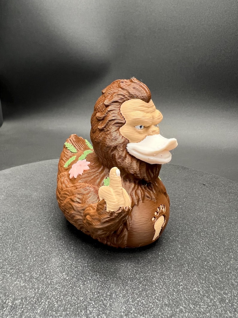 Puede incluir: Una figura con forma de pato, marr&oacute;n y peluda, con cara de color tostado claro y pico blanco. La figura tiene una mano levantada haciendo un gesto con el dedo coraz&oacute;n, un emblema de huella en el pecho y peque&ntilde;as flores rosas y hojas verdes.