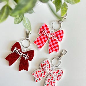 May include: Three bow-shaped keychains with names. One is red glitter, another is red and pink checkered, and the third is white with red cherries. Each has a silver clasp and a name in white script: Blakely, Aubree, and Charlotte.