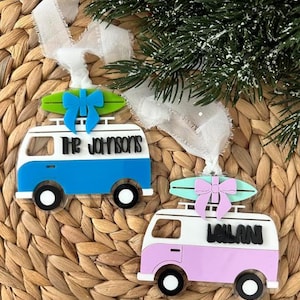 May include: Two decorative ornaments shaped like vintage camper vans. One is blue and white with a green surfboard and a blue bow, the other is pink and white with a green surfboard and a pink bow. Both have black wheels and the family name on the side.