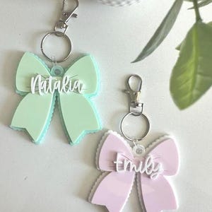 May include: Two bow-shaped keychains with silver-toned metal lobster clasps and rings. One keychain is mint green with the white script name Natalia, and the other is light pink with the white script name emily attached to the center.