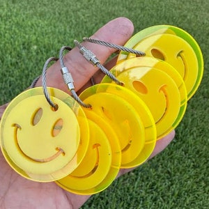 May include: A collection of yellow smiley face keychains. Each keychain features a circular design with a smiling face cutout and a silver metal loop for attaching keys. The keychains are held in a hand against a green grassy background.