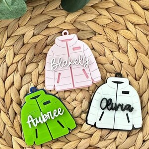 May include: Three colorful jacket-shaped ornaments. One is light pink with the name "Blakely", another is green with "Aubree", and the last is white with "Olivia". Each has a loop for hanging and a zipper detail.