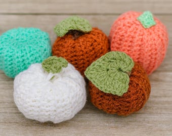 May include: A collection of five crocheted pumpkins in various colors: white, orange, coral, and teal. Each pumpkin has a green crocheted leaf and stem, perfect for fall decor. The pumpkins are arranged on a wooden surface.