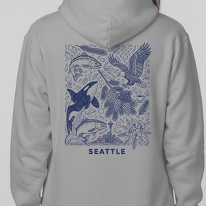 May include: A light gray hoodie with a blue graphic on the back featuring marine life and an eagle. The word "SEATTLE" is printed in blue at the bottom of the design. The hoodie has a hood and long sleeves.