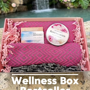 Könnte beinhalten: Eine Geschenkbox mit einem gefalteten, magenta- und graufarbenen Textil mit Muster, einer runden Dose mit der Aufschrift Rosengarten und einer Visitenkarte der Amla Mecklenburgische Seifenmanufaktur. Die Box enthält rosa Füllmaterial auf Stein.