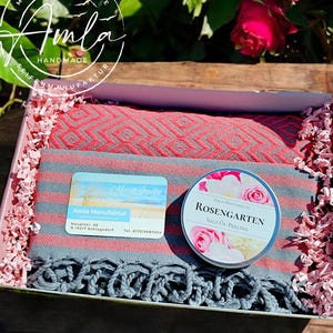 Könnte beinhalten: Eine Geschenkbox mit einem grau-rot gestreiften Textil mit Fransen, einem rot-grauen Textil mit Rautenmuster und einer runden Dose mit der Aufschrift Rosengarten Salz-Öl-Peeling und Rosenmotiv, umgeben von rosa Papierstreifen.