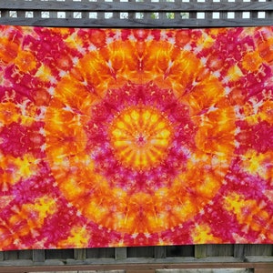 May include: A large, circular tie-dye tapestry with a mandala design in shades of orange, yellow, and pink. The tapestry is hanging on a wooden fence.
