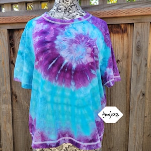 May include: A tie-dye t-shirt in shades of blue and purple, with a spiral design at the centre. The shirt has short sleeves and a round neckline. The brand name "Amy Lora" is visible on the lower right.